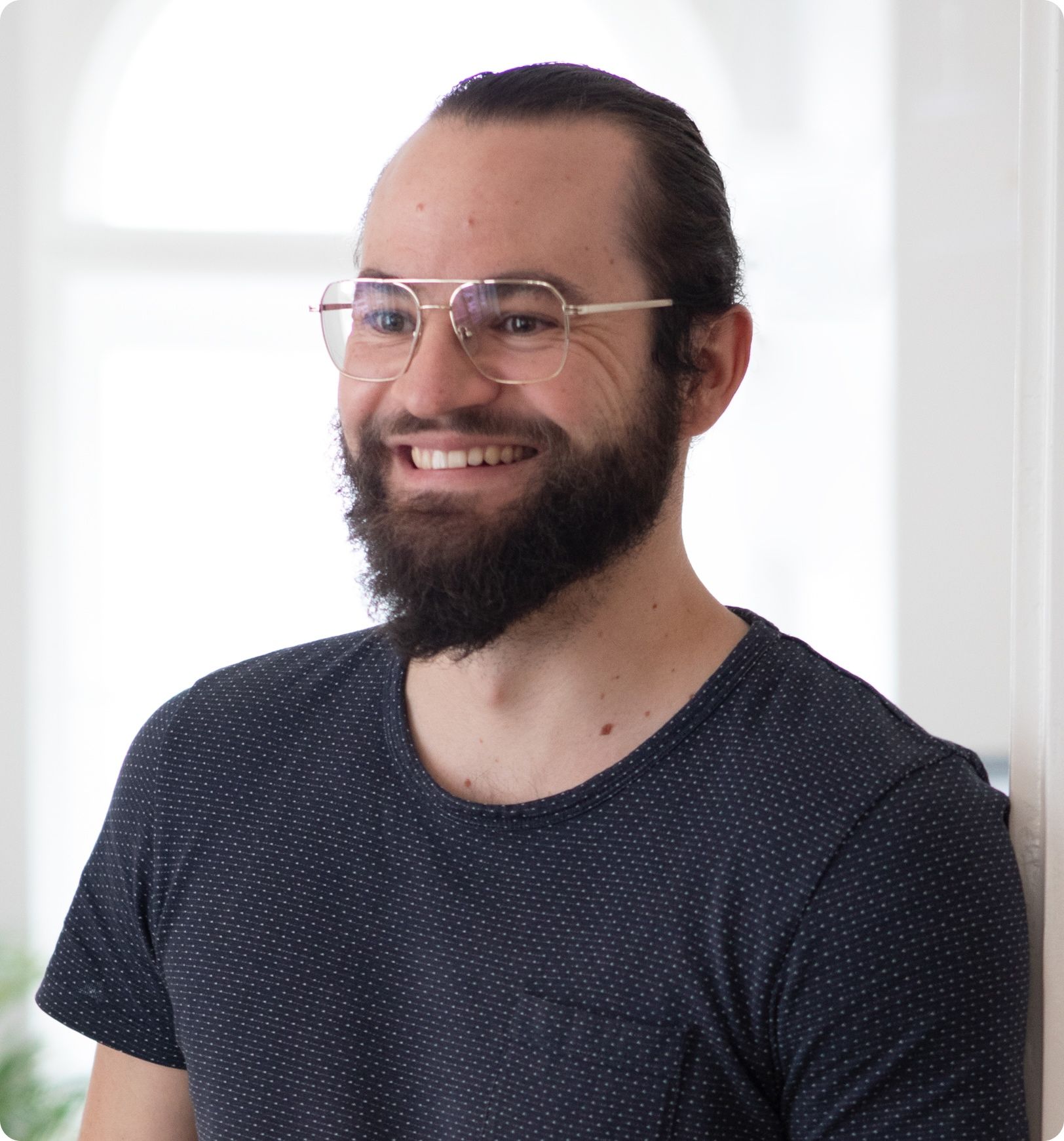 Valentin, smiling with glasses, beard and a dotted navy t-shirt by a window, loomobox team
