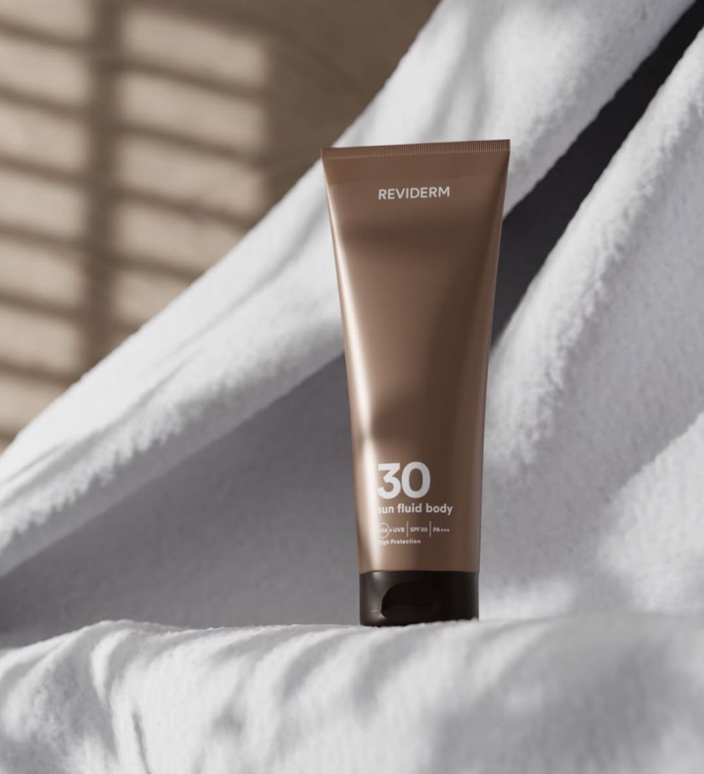 REVIDERM sun fluid body SPF 30 tube standing on white towels in striped sunlight